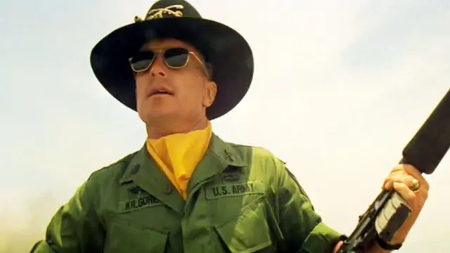 روبرت دوفال في مشهد من "القيامة الآن" بزي عسكري كتب عليه يو أس أرمي - الجيش الأمريكي - مع قبعة عسكرية وبندقية في يديه اليمنى