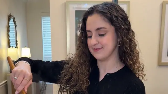 تظهر في الصورة امرأة ترتدي قميصاً أسود، تقف أمام منضدة مطبخ وتنظر إلى الأسفل بينما تحرّك الطعام بملعقة خشبية، فيما تنسدل خصلات شعرها المجعّد بلون بني على كتفيها.