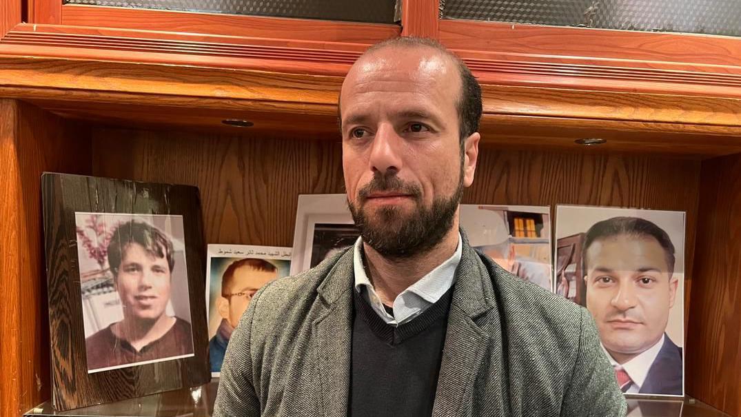 المحامي فادي العبار، ممثل رابطة عائلات قيصر في دمشق تظهر خلفه مجموعة صور.