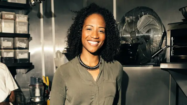 تبتسم امرأة ذات شعر أسود مجعّد للكاميرا داخل مطبخ، وتقف إلى جانب مروحة سوداء كبيرة. وتظهر خلفها أدوات مطبخ، إضافة إلى ذراع رجل يبدو أنه يعمل في الخلفية.
