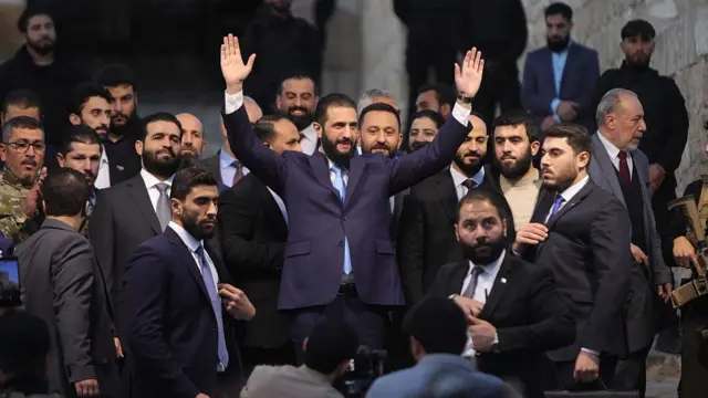 الرئيس أحمد الشرع، مرتدياً بذلة زرقاء داكنة وربطة عنق زرقاء، رافعاً يديه في الهواء تحيّة للجماهير، خلال فعالية أقيمت في نوفمبر/تشرين الثاني 2025 للاحتفال بذكرى انتهاء سيطرة نظام الأسد على حلب. ويتميز الشرع بشعر رأس ولحية داكنين، ويحيط به رجال آخرون يرتدون بذلات رسمية.