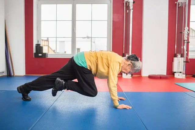 أمرأة مُسنة، ترتدي سروالاً رياضياً وحذاءً أسود رياضياً ونظارة نظر وسترة صفراء رياضية، وتمارس التمارين الرياضية في صالة ألعاب رياضية.