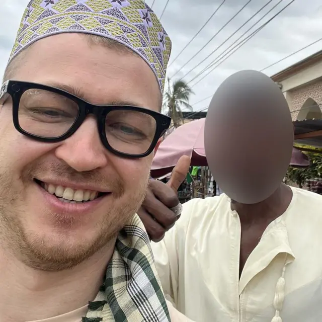 رجل يرتدي نظارات شفافة بإطار داكن من علامة راي بان، تظهر فيها كاميرا صغيرة فوق العدسة اليسرى. كما يرتدي قبعة تقليدية مزخرفة بنمط ماسي باللونين الأصفر والبنفسجي.