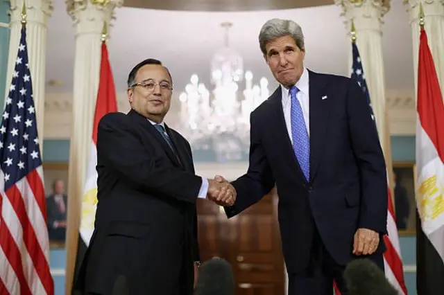 كيري يرتدي بدلة رسمية كحلية اللون وقميصاً أبيض وربطة عنق زرقاء يقف إلى يمين الصورة مصافحاً فهمي الذي يرتدي بدلة رسمية سوداء وقميصاً أبيض اللون وربطة عنق بلون فيروزي. وخلفهما أعلام مصر والولايات المتحدة.