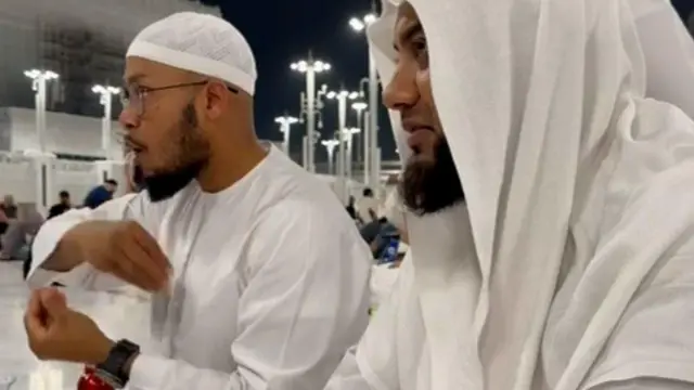 رجلان يرتديان ملابس إسلامية بيضاء يجلسان في فضاء معماري واسع ومضيء، تتخلله أعمدة عالية وأضواء في الخلفية، ويتواصلان باستخدام لغة الإشارة. ويستخدم أحدهما، الجالس على اليسار، لغة الإشارة البريطانية.