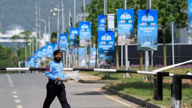 حارس أمن يسير في طريق خالٍ، وتتدلى من أعمدة الإنارة ملصقات زرقاء تحمل عبارة "محادثات إسلام آباد" باللون الأبيض