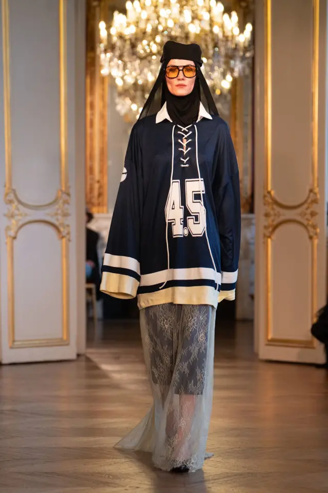 عارضة أزياء ترتدي قميصاً رياضياً طويلاً، وتنورة، وغطاء رأس، ونظارة شمسية ذات عدسات صفراء، وقبعة.
