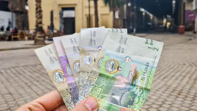 صورة ليد رجل تحمل دنانير كويتية ويبرز في خلفية الصورة شارع.