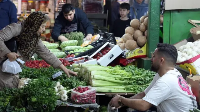 امرأة تظهر من الجانب يبدو أنها كبيرة في السن، ترتدي على رأسها شالاً منقطاً لونه كلون جلد النمر، مع سترة طويلة لونها بني فاتح، تميل لتلتقط بيدها خضراوات من مكان يعرض الخضراوات، وهناك رجل يجلس قرب المكان أمامها، وعدد آخر من الأشخاص خلفها.
