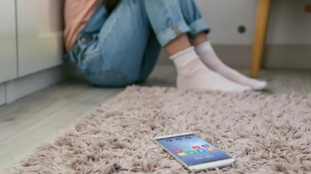 صورة لهاتف خلوي ملقى على الأرض وفي خلفية الصورة فتاة تجلس أرضا في الزاوية.