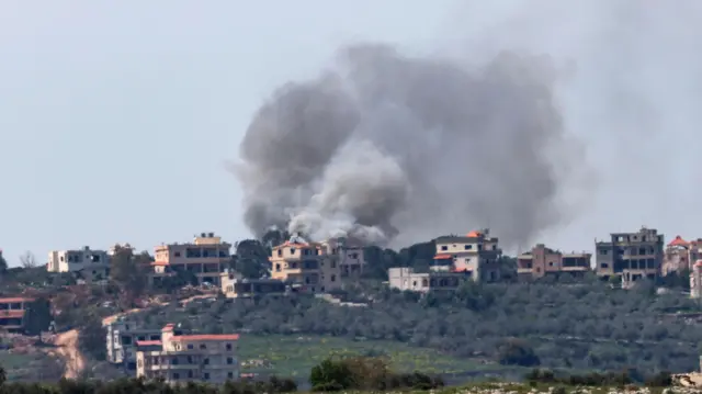 تلة تظهر فيها عدة منازل ودخان قصف وأشجار زيتون - صورة من بلدة بنت جبيل