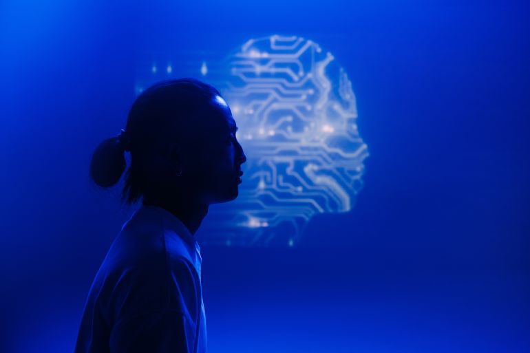 A human silhouette against a digital projection of a head, resembling a blue-lit circuit. The futuristic scene symbolizes artificial intelligence, technology, and the fusion of man and machine.