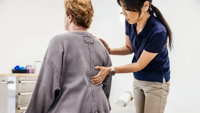 مقدمة رعاية صحية تضع يدها على ظهر امرأة أثناء فحصها، فيما تجلس المرأة داخل غرفة في عيادة.
