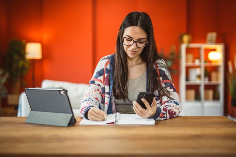 adult woman work from home use mobile phone and write on notebook