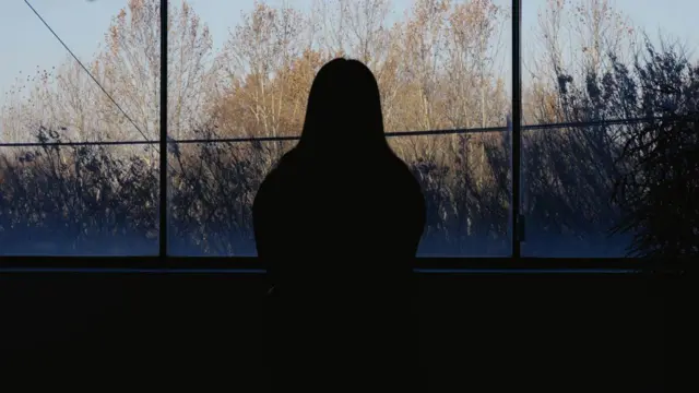 صورة ظلية لامرأة تنظر من نافذة، وتظهر في الخلفية أشجار ونباتات.
