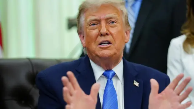 الرئيس الأمريكي دونالد ترامب يشير بيديه أثناء حديثه مع الصحفيين في المكتب البيضاوي للبيت الأبيض. ويرتدي بدلة رسمية كحلية اللون مع ربطة عنق بتدرجات اللون الأزرق وقميص أبيض.