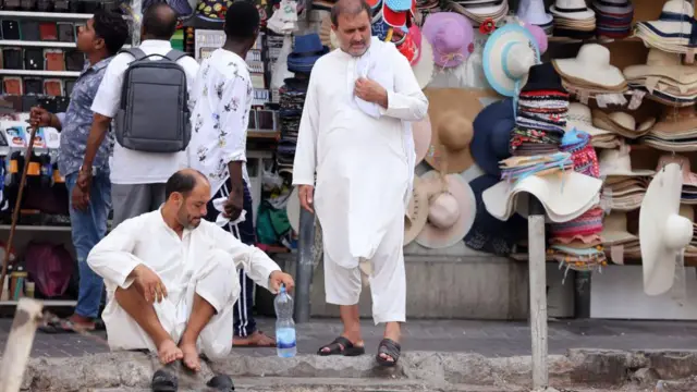 رجلان بعباءة بيضاء أحدهما جالس يمسك بقارورة ماء وآخر يقف بجانبه وخلفهما بائع قبعات وزبونان