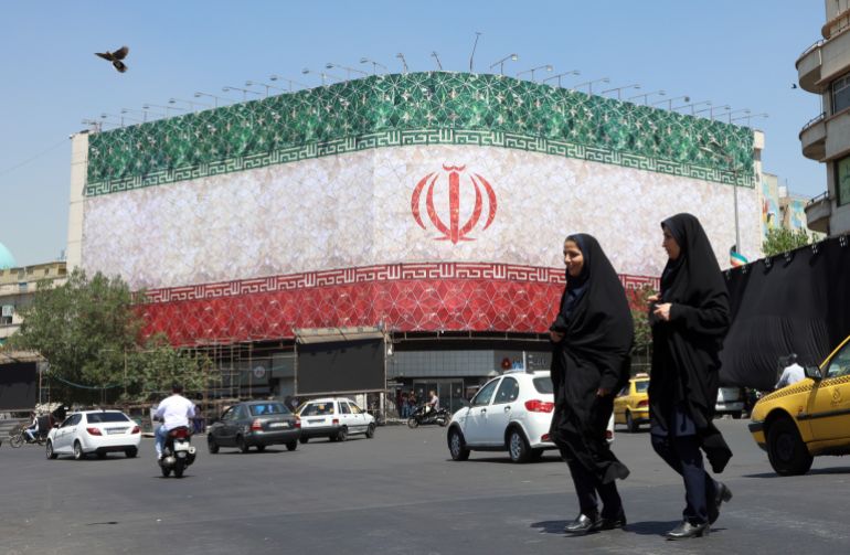 epa13035937 Veiled Iranian women walk past a billboard featuring Iran's national flag at Enqelab Square in Tehran, Iran, 14 June 2026. US President Donald Trump saying on a social media platform that a deal could be signed soon. EPA/ABEDIN TAHERKENAREH RESTRICTIONS: NO Access Israel Media/Persian Language TV Stations Outside Iran/Strictly No Access BBC Persian/VOA Persian/Manoto TV/Iran International TV. (As mandated by Iran's Directorate General for Foreign Media) --