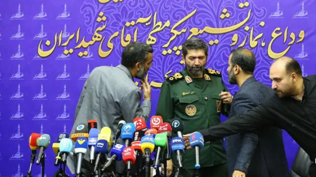 يتحدث قائد فيلق محمد رسول الله، حسن حسن زاده، مرتدياً زيه العسكري، إلى رجلين بملابس مدنية، بينما يضبط رجل ثالث مجموعة من الميكروفونات الملوّنة خلال مؤتمر صحفي.