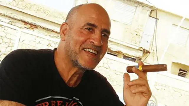 دانيال سياد، رجل حليق الرأس ولديه لحية خفيفة مرتدياً قميصاً أسود اللون، يجلس في الخارج فيما يبدو أنه شارع، مبتسماً ويرفع سيجاراً كبيراً في يده.