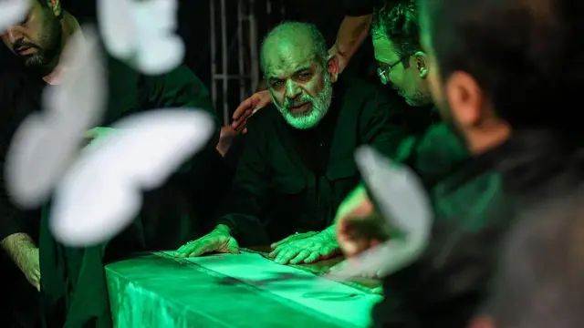 رجل بلحية بيضاء وملابس سوداء يضع يده على نعش مغطى بعلم إيران
