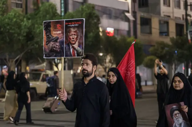 شاب يرتدي قميصاً أسود وشعره أسود كثيف، ولديه لحية سوداء خفيفة تغطي وجهه، بينما يحمل لافتة مناهضة للولايات المتحدة لدى وصوله مع آخرين لحضور مراسم تشييع الزعيم الإيراني الراحل علي خامنئي وأفراد من عائلته بالقرب من مرقد الإمام الرضا في مدينة مشهد، في وقت مبكر من يوم 9 يوليو/تموز عام 2026. ويستعد الإيرانيون اليوم لدفن زعيمهم الراحل علي خامنئي في مسقط رأسه بمدينة مشهد، شمال شرق إيران وتأتي مراسم دفن المرشد الأعلى الراحل عقب جنازة استمرت عدة أيام وجذبت ملايين المشيعين في مدن مختلفة بإيران والعراق.