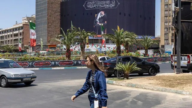 امرأة بملابس زرقاء ونظارة شمسية تعبر شارعا في طهران وخلفها صورة عملاقة لعلي خامنئي باللون الأسود