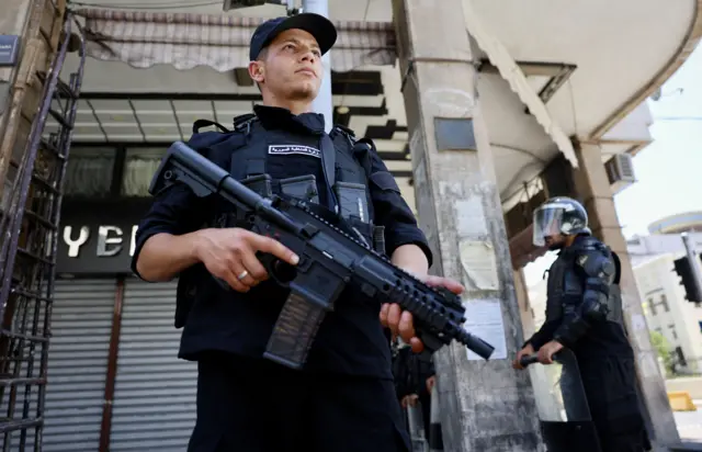 يظهر في الصورة أحد عناصر الأمن السوري أمام مبنى البرلمان في دمشق، حيث يقف مدججاً بالسلاح، ويرتدي قبعة سوداء وزياً شرطياً أسود.