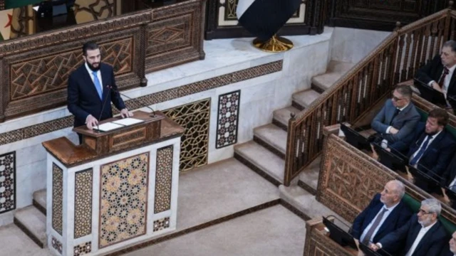  الرئيس السوري المؤقت أحمد الشرع، يلقي كلمة خلال جلسة مجلس الشعب الأولى منذ سقوط الأسد، وذلك في العاصمة السورية دمشق، ويرتدي الشرع سترة سوداء وقميصاً أبيض وربطة عنق لونها أزرق فاتح، بينما يقف متحدثاً من على منبر ويجلس أمامه أعضاء مجلس الشعب.