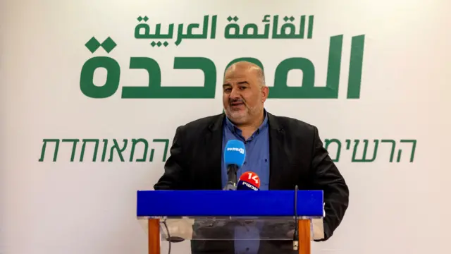 صورة لرجل (منصور عباس) يتحدث أثناء اجتماع لحزبه في الكنيسيت، وقد كتب وراءه: "القائمة العربية الموحدة" باللغتين العربية والعبرية